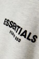 Close-up of the gray fabric on the Fear of God Essentials Long Sleeve Polo Sweatshirt, featuring black embroidered text reading "ESSENTIALS FEAR OF GOD.