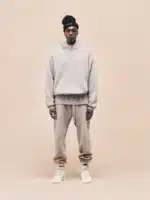 A person stands against a beige background wearing a beige hooded sweatshirt, Fear of God Sweatpant 'Core-Heather', white sneakers, and a black beanie. They face forward with a neutral expression.