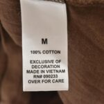 Close-up of a clothing label on a brown fabric. The label reads: M, 100% Cotton, Exclusive of Decoration, Made in Vietnam, RN# 090233, Over for Care. Product: Fear of God Essentials Knitted Sweater Harvest.