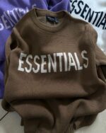 Three Fear of God Essentials Knitted Sweaters are displayed, each prominently featuring the word "ESSENTIALS" on the front. The sweaters are shown in brown, white, and purple hues, neatly folded and layered atop one another on a flat surface.