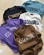 Five Fear of God Essentials Knitted Sweaters with "ESSENTIALS" printed on them are laid out on a tiled floor. The colors are black, teal, brown, purple, and white.