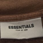 A label displaying the product name "Fear of God Essentials Knitted Sweater Harvest" with black text reading ESSENTIALS and FEAR OF GOD on a beige background is attached to the brown fabric of the sweater.