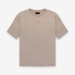 A Fear of God Essentials FG T-shirt in 'Dusty-Beige' featuring short sleeves and a minimalist design. The letters FG are subtly printed on the chest in a slightly darker hue. The t-shirt is displayed flat against a plain white background.