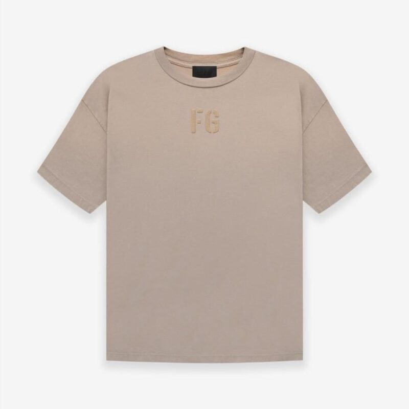 A Fear of God Essentials FG T-shirt in 'Dusty-Beige' featuring short sleeves and a minimalist design. The letters FG are subtly printed on the chest in a slightly darker hue. The t-shirt is displayed flat against a plain white background.