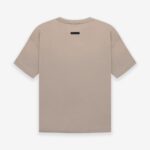 A Fear of God Essentials FG T-shirt in 'Dusty-Beige' is shown from the back, showcasing a plain design with short sleeves and a small black rectangular label near the neckline, all against a white background.