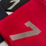 A close-up showcases two sports jerseys on a surface, one black and the other a prominently displayed red, each adorned with the number 7 in white. The red jersey is labeled as the Fear of God Essentials 7 T-Shirt 'Red'.