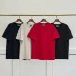 Four Fear of God Essentials 7 T-Shirts in various colors—black, beige, red, and black—are displayed on wooden hangers on a metal rod against a simple white background.