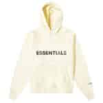 The Fear of God Essential Hoodie in 'Buttercream' is cream-colored with bold black ESSENTIALS lettering on the front. It includes a front pocket and hood, with smaller ESSENTIALS text printed on the left sleeve near the wrist.