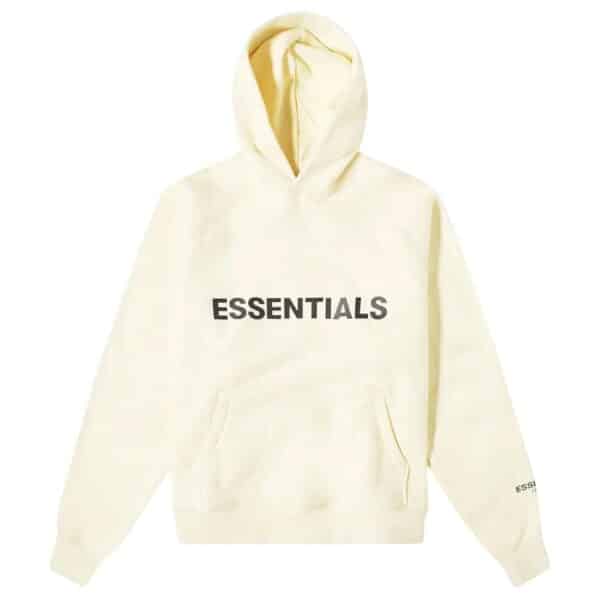 The Fear of God Essential Hoodie in 'Buttercream' is cream-colored with bold black ESSENTIALS lettering on the front. It includes a front pocket and hood, with smaller ESSENTIALS text printed on the left sleeve near the wrist.