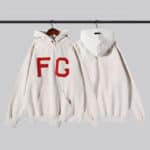 Two Fear of God Essentials Hoodies in Cream-Heather hang on a clothing rack. The front of one hoodie displays large red letters "FG," while the back of the other hoodie is plain. Both hoodies feature front pockets and ribbed cuffs.