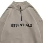 A beige Essentials Half Zip High Collar Loose Hoodie with "ESSENTIALS" printed in bold black letters across the chest, featuring a high collar and a simple, casual design.