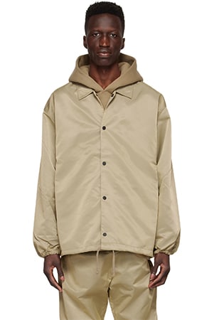 A person wearing the Essentials Tan Nylon Jacket and matching pants stands against a white background. The jacket, featuring buttons down the front and an oversized fit, has its hood up, partially covering the person's head.