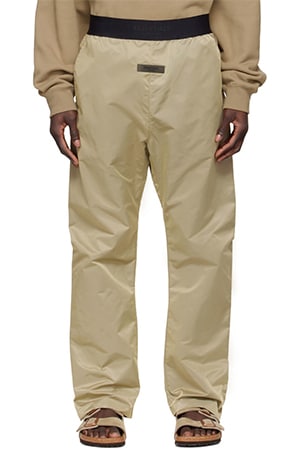 A person is standing on a plain white background wearing the Essentials Tan Nylon Lounge Pants, which feature an elastic waistband and a small logo patch at the front. They are also wearing a beige hoodie and brown sandals.