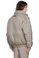 A person with long hair is wearing an Essentials Taupe Polyester Jacket, featuring a high collar and gathered cuffs. The jacket has an oversized fit, appears quilted for warmth, and is viewed from the back with a slight turn to the side.