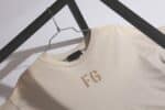 A Dusty-Beige Fear of God Essentials FG T-shirt is draped on a metal hanger, showcasing the subtle matching FG letters on its front, all set against a geometric black metal frame.