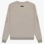 The FG Essentials Crewneck Sweatshirt 'Core-Heather' is showcased on a white background. It boasts a light beige color with long sleeves adorned with subtle G letters on each arm. The sweatshirt's minimalistic design is complemented by a small black tag near the back neckline.