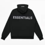 The Fear Of God Essentials Reflective Letter Hoodie, in black, is displayed flat on a white background. It prominently features the word ESSENTIALS printed in large white letters across the back, with the hood visible at the top.