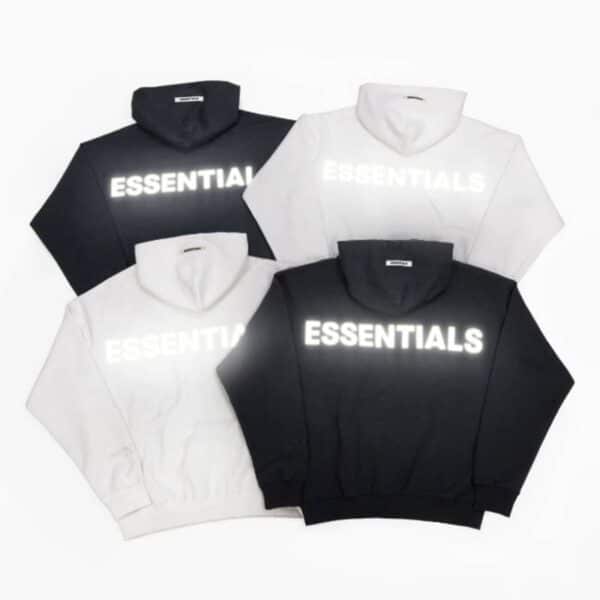 A square arrangement featuring four Fear Of God Essentials Reflective Letter Hoodies, consisting of two black and two white, each showcasing the word ESSENTIALS in bold, reflective print on the back. The background is a plain white canvas.