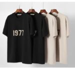 Displayed on wooden hangers against a white background are four minimalist Fear of God Essentials 1977 T-shirts. Two black shirts prominently feature the '1977' text in white, while two beige shirts with the same text pattern are partially visible.