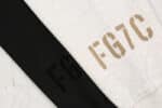 Close-up of black and white fabric swatches. The white fabric has the letters 'Fear of God Essentials FG7C' printed in bold brown text, while the black fabric has the letters 'FG' printed in bold black text. Both fabrics are placed on a light gray surface.