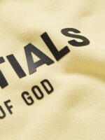 A close-up of the Fear Of God Blend Jersey Essentials Hoodie in beige, featuring the word "ESSENTIALS" in bold black letters prominently displayed, with smaller text partially visible beneath it. The texture of the fabric is clearly evident.